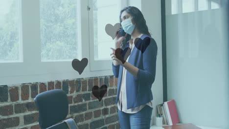 Animation-of-hearts-floating-over-biracial-woman-in-face-mask-having-call-in-office