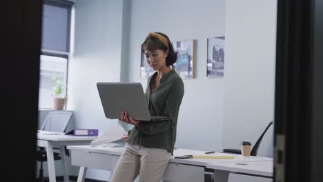 Biracial-businesswoman-standing-at-table-and-using-laptop-alone-at-office