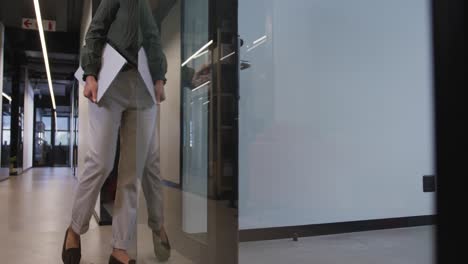 Biracial-businesswoman-walking-with-laptop-and-opening-door-at-office