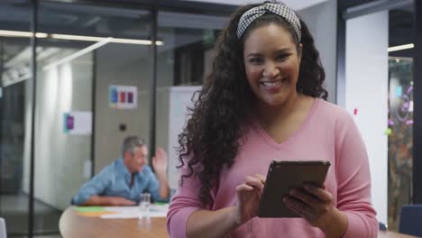 Portrait-of-happy-biracial-businesswoman-using-tablet-and-looking-at-camera-at-office