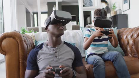 Video-Eines-Glücklichen-Afroamerikanischen-Vaters-Und-Sohnes,-Der-Auf-Dem-Sofa-Sitzt-Und-Mit-Dem-VR-Headset-Spiele-Spielt