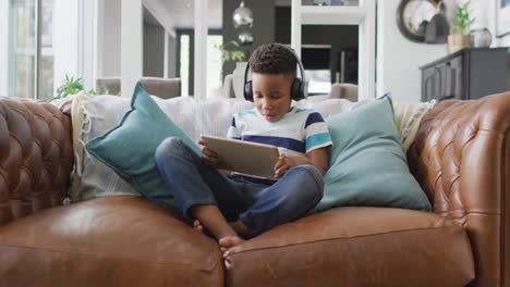 Video-Eines-Afroamerikanischen-Jungen-Mit-Kopfhörern,-Der-Auf-Dem-Sofa-Sitzt-Und-Ein-Tablet-Benutzt
