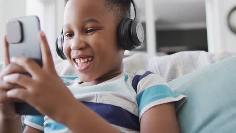 Video-Eines-Glücklichen-Afroamerikanischen-Jungen-Mit-Kopfhörern,-Der-Auf-Dem-Sofa-Sitzt-Und-Ein-Tablet-Benutzt