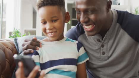 Video-of-happy-african-american-father-and-son-sitting-on-sofa-and-using-smartphone