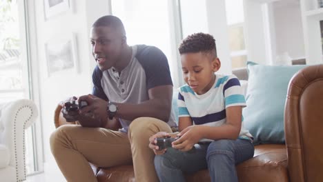 Video-Eines-Glücklichen-Afroamerikanischen-Vaters-Und-Sohnes,-Der-Auf-Dem-Sofa-Sitzt-Und-Videospiele-Spielt