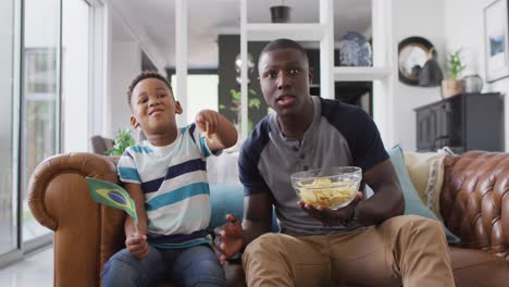 Video-Eines-Glücklichen-Afroamerikanischen-Vaters-Und-Sohnes,-Der-Auf-Dem-Sofa-Sitzt-Und-Sich-Ein-Spiel-Im-Fernsehen-Ansieht