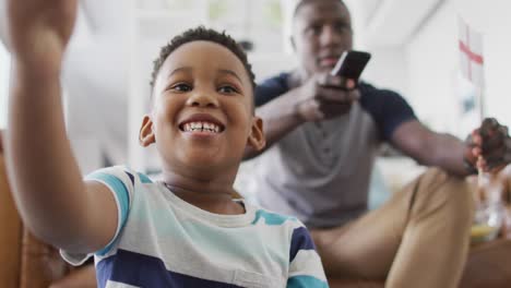 Video-of-happy-african-american-father-and-son-sitting-on-sofa-and-watching-match-in-tv