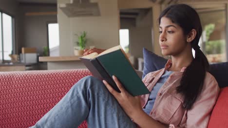 Video-of-biracial-woman-sitting-on-sofa-and-reading-book