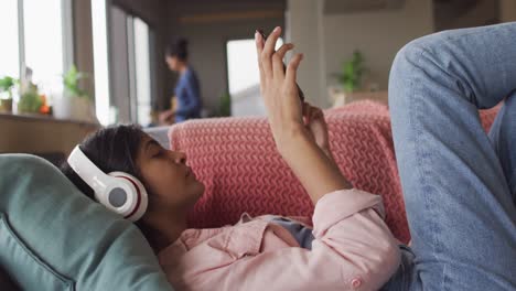 Video-Einer-Entspannten-Gemischtrassigen-Frau-Mit-Kopfhörern,-Die-Auf-Dem-Sofa-Liegt-Und-Ihr-Smartphone-Benutzt