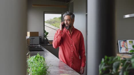 Biracial-man-wearing-red-shirt-talking-on-smartphone-in-kitchen-alone