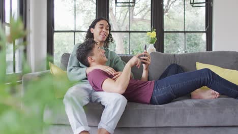 Happy-diverse-male-couple-sitting-on-sofa,-embracing-and-using-smartphone-in-living-room
