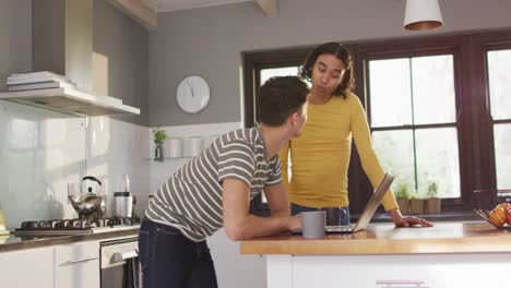 Happy-diverse-male-couple-drinking-coffee-and-using-laptop-in-kitchen
