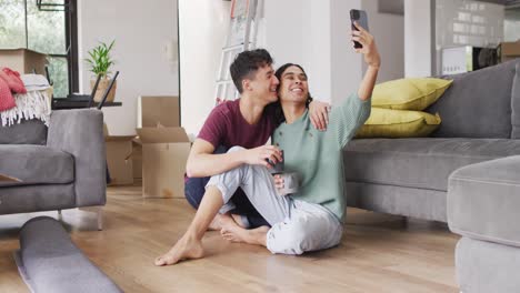 Happy-diverse-male-couple-moving-house,-drinking-coffee,-taking-selfie-using-smartphone