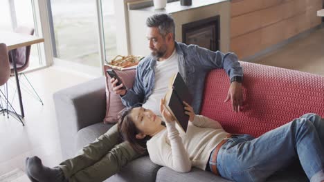 Happy-diverse-couple-sitting-in-living-room-with-book-and-smartphone