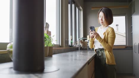 Asian-woman-wearing-jumper-and-using-smartphone-in-kitchen-alone