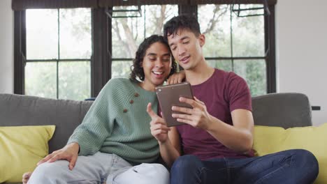 Happy-diverse-male-couple-sitting-on-sofa,-embracing-and-using-tablet-in-living-room