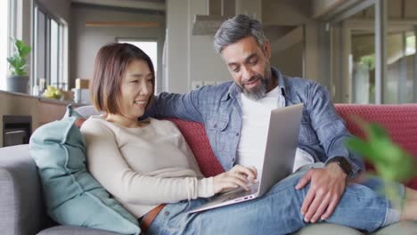 Happy-diverse-couple-sitting-in-living-room-using-laptop-and-talking