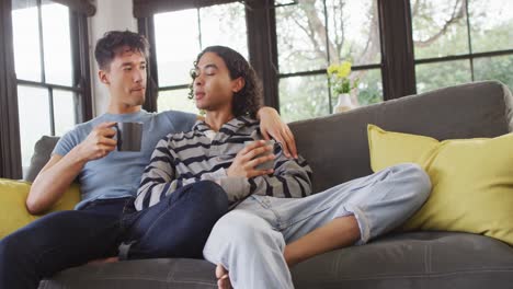 Happy-diverse-male-couple-sitting-on-sofa-and-drinking-coffee-in-living-room