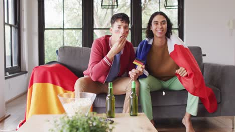 Happy-diverse-male-couple-with-national-flags-watching-tv-in-living-room