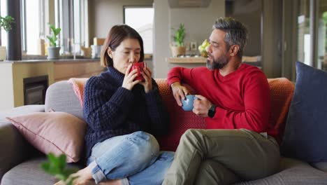 Happy-diverse-couple-sitting-in-living-room-drinking-coffee-and-talking
