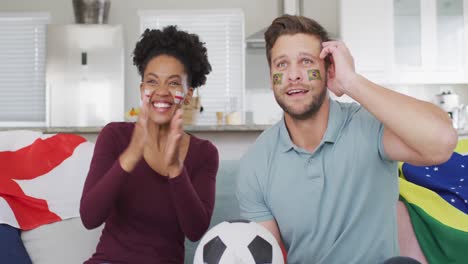 Video-of-happy-diverse-couple-with-flags-of-portugal-and-england-and-watching-match-in-tv