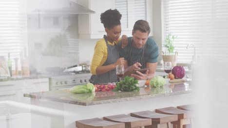 Video-of-happy-diverse-couple-preparing-meal,-using-tablet-and-drinking-wine-in-kitchen