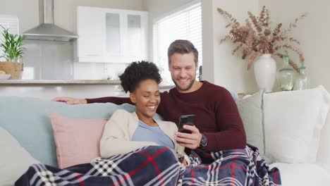 Video-of-happy-diverse-couple-embracing-and-using-smartphone-on-sofa