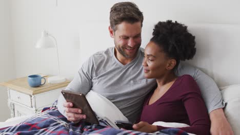 Video-Eines-Glücklichen,-Vielfältigen-Paares,-Das-Im-Bett-Ein-Tablet-Benutzt-Und-Spricht