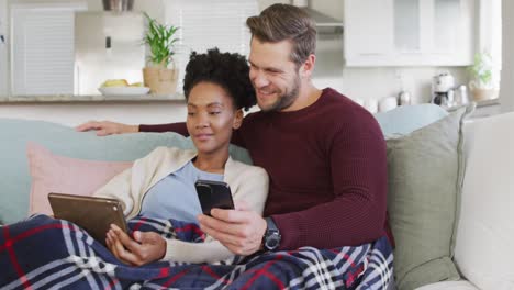 Video-of-happy-diverse-couple-embracing-and-using-electronic-devices-on-sofa
