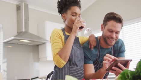 Video-of-happy-diverse-couple-preparing-meal,-using-tablet-and-drinking-wine-in-kitchen
