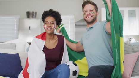Video-of-happy-diverse-couple-holding-flags-of-portugal-and-england-and-watching-match-in-tv