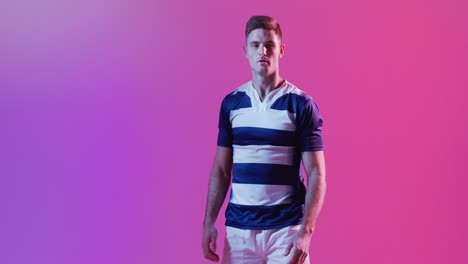 Portrait-of-caucasian-male-rugby-player-with-rugby-ball-over-pink-lighting