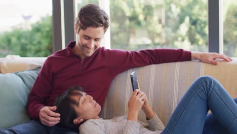 Video-of-back-view-of-happy-diverse-couple-sitting-on-sofa-with-smartphone
