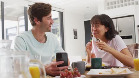 Video-of-happy-diverse-couple-using-smartphone-and-drinking-coffee-together-in-kitchen
