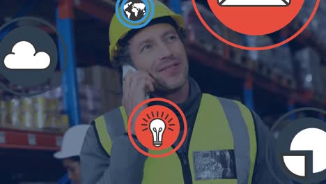 Animation-of-icons-against-caucasian-factory-worker-in-protective-wear-talking-on-phone-in-warehouse