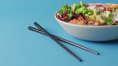 Composition-of-bowl-of-rice-and-vegetables-with-chopsticks-on-blue-background