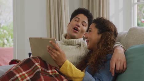 Video-of-happy-biracial-couple-siting-on-sofa-under-blanket-and-using-tablet