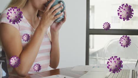 Animation-of-coronavirus-cells-over-caucasian-young-woman-drinking-coffee-while-working-at-desk