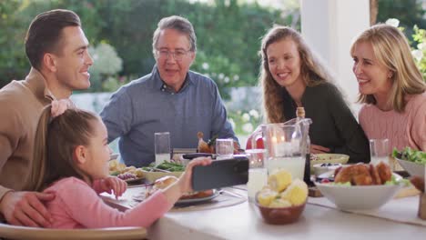 Video-of-happy-caucasian-parents,-daughter-and-grandparents-at-dinner-table-looking-at-smartphone