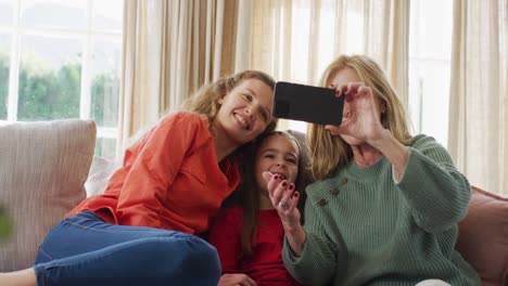 Video-of-happy-caucasian-mother-and-grandmother-looking-at-smartphone-with-granddaughter-on-couch