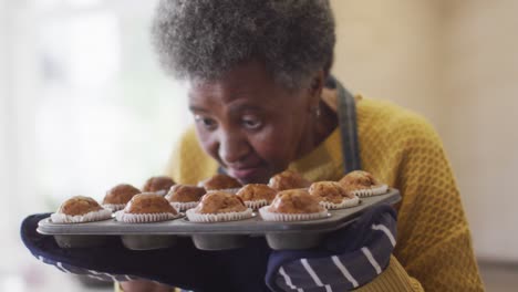 Video-Einer-Glücklichen-älteren-Afroamerikanerin,-Die-Ein-Tablett-Mit-Frisch-Gebackenen-Cupcakes-In-Der-Hand-Hält-Und-Duftet