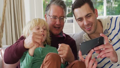 Video-of-happy-caucasian-father,-grandfather-and-grandson-sitting-on-couch-looking-at-smartphone