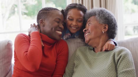 Video-of-smiling-african-american-mother,-daughter-and-grandmother-sitting-hugging-in-living-room