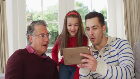 Video-of-happy-caucasian-father,-grandfather-and-granddaughter-sitting-on-couch-looking-at-tablet