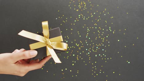 Woman's-hand-holding-black-gift-box-with-gold-ribbon,-over-glitter-on-black-with-copy-space