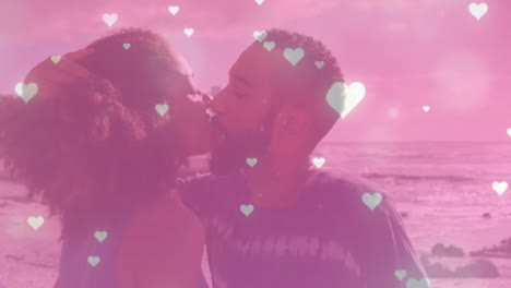 Animation-of-glowing-pink-heart-icons-falling-over-african-american-couple-kissing-at-the-beach