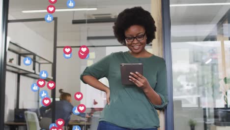 Animation-of-social-media-icons-over-happy-african-american-woman-using-tablet-in-office