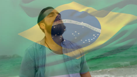 Zusammengesetztes-Video,-In-Dem-Die-Brasilianische-Flagge-über-Einem-Kaukasischen-Mann-Mit-Geschlossenen-Augen-Schwenkt,-Der-Am-Strand-Steht