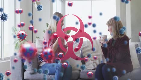 Animation-of-virus-cells-and-biohazard-symbol-over-caucasian-female-friends-sitting-on-sofa