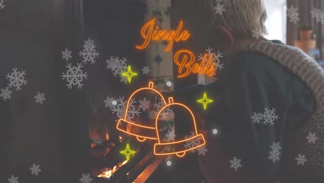 Animation-of-jingle-bells-text-in-red-neon,-christmas-bells-and-snowflakes-over-boy-lighting-fire
